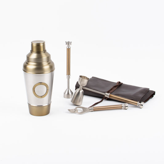 A cocktail kit consisting of a steel jigger, muddler, and bottle opener alongside a brass cocktail shaker and a brown pouch. All items have a metallic finish with brass accents.