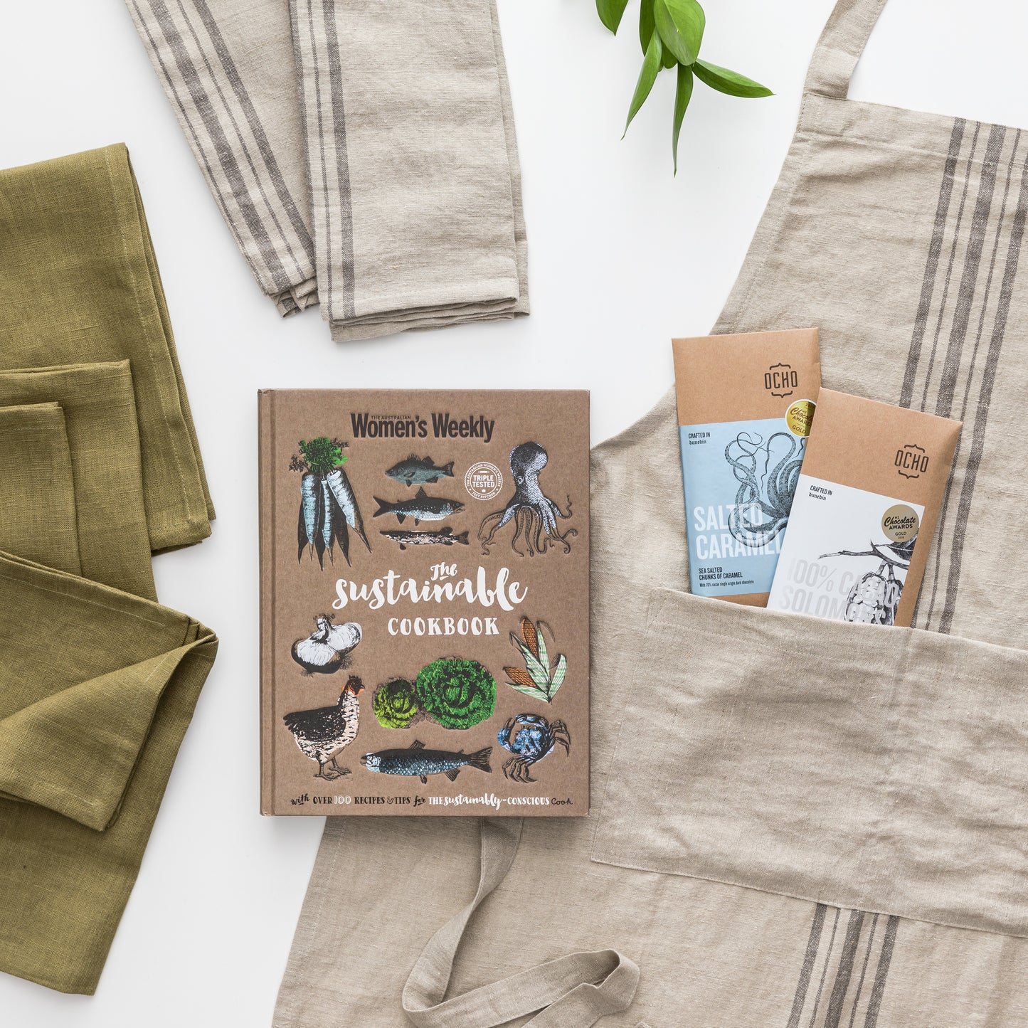 Flatlay of products are apron, two tea towels, four linen napkins, cookbook, two chocolates.
