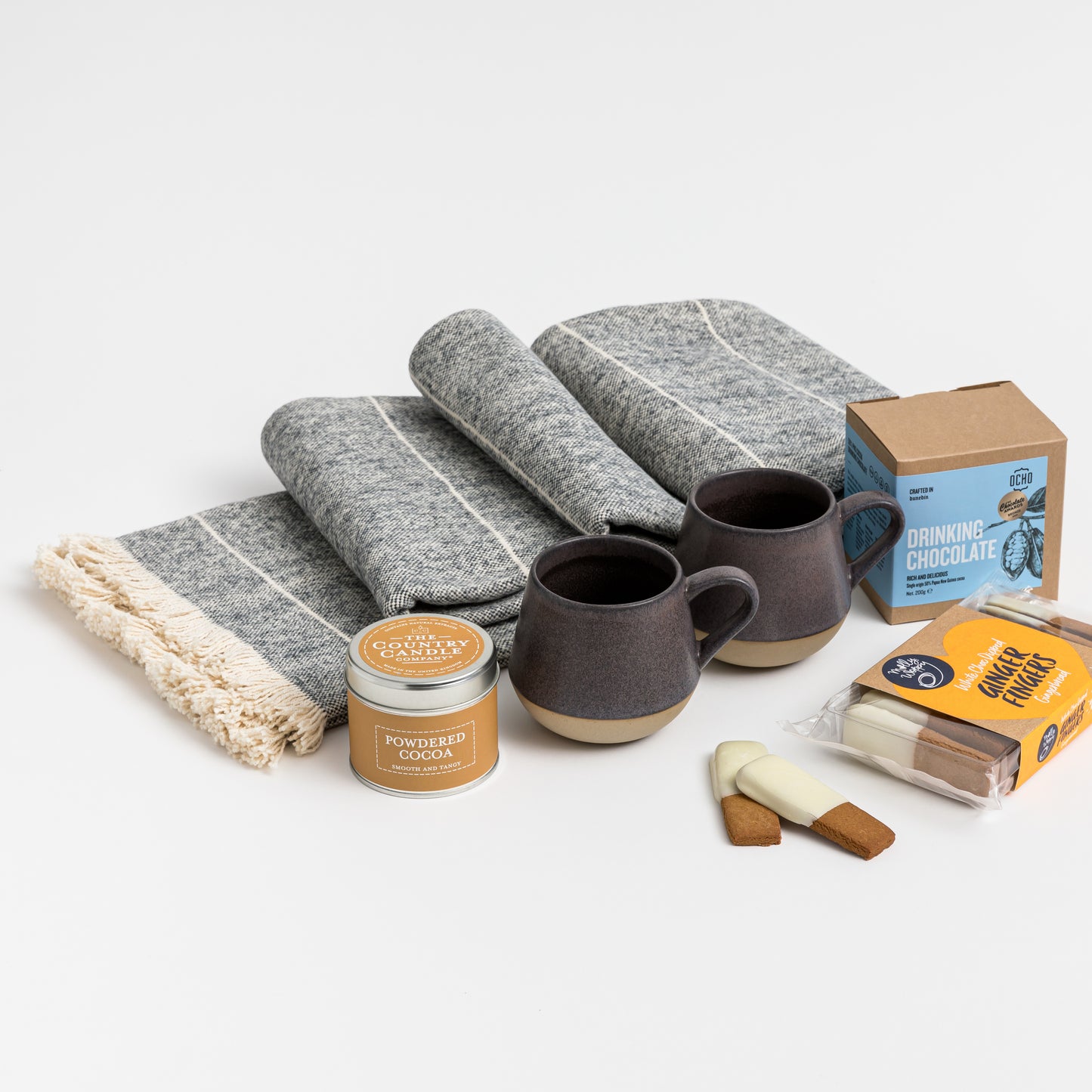 Flatlay of products out of gift box are cosy throw, two ceramic mugs, biscuits, hot chocolate, candle.