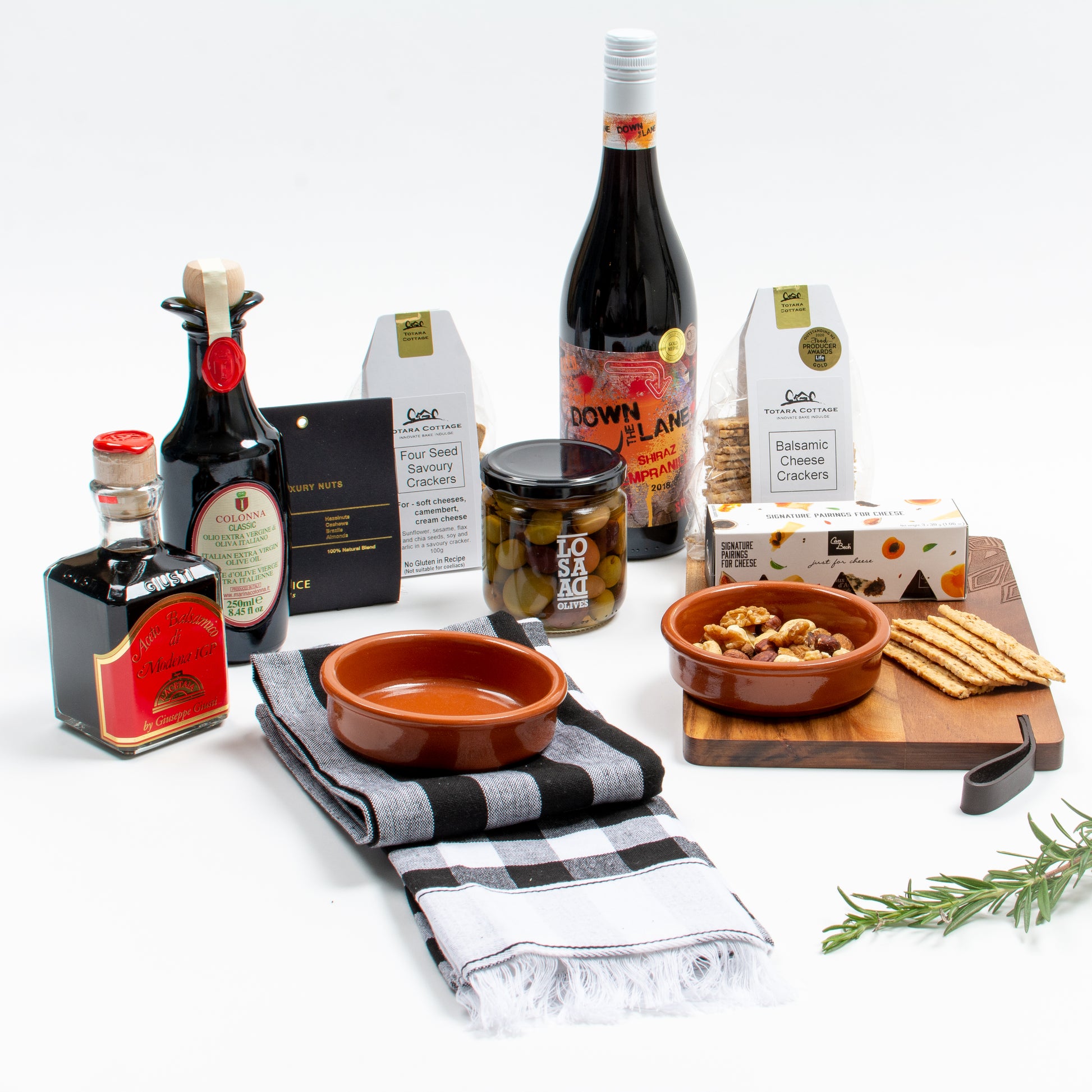 Products displayed out of gift box are wooden board, two tea towels, two tapas dishes, two crackers, nuts, cheese jellies, olives, olive oil, balsamic vinegar, red wine.